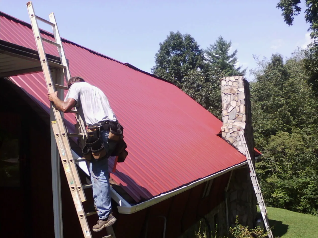 Roofer on a ladder beside a red metal roof and stone chimney — Soffit & Fascia Repair by Roofing Brothers in Sulligent